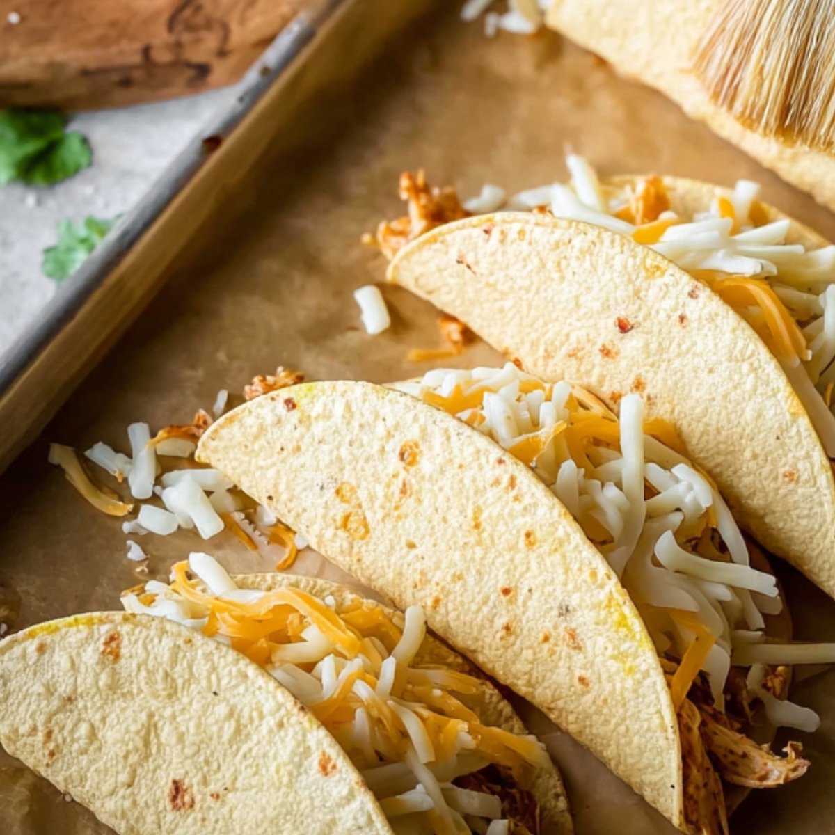 Top-down view of four soft tacos on a parchment-lined baking sheet, filled with shredded chicken and shredded cheese, with a hand brushing oil on one taco using a pastry brush.