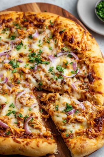 A freshly baked BBQ chicken pizza on a wooden cutting board, topped with shredded chicken, melted cheese, red onion slices, and chopped parsley. One slice is slightly pulled out, and a small dish of extra parsley is visible in the background.