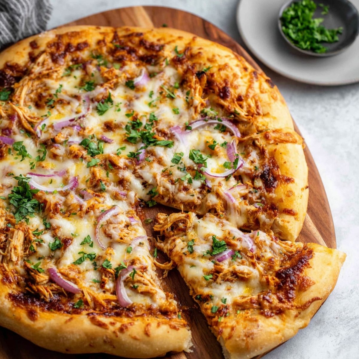A freshly baked BBQ chicken pizza on a wooden cutting board, topped with shredded chicken, melted cheese, red onion slices, and chopped parsley. One slice is slightly pulled out, and a small dish of extra parsley is visible in the background.
