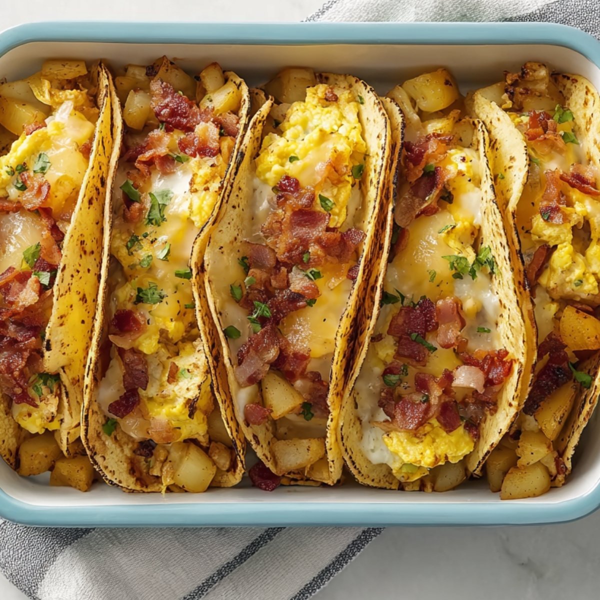A rectangular baking dish with five baked breakfast tacos, each filled with scrambled eggs, diced cooked potatoes, crispy bacon, and melted cheese, garnished with chopped parsley.