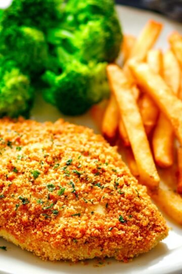 A crispy baked ranch chicken breast served on a white plate with golden French fries and steamed broccoli, garnished with herbs, showing a finished meal.