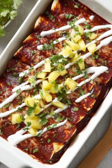 Close-up of a white rectangular baking dish with beef and pineapple enchiladas, covered in smooth red enchilada sauce, drizzled with sour cream and sprinkled with fresh cilantro and pineapple chunks, bright natural lighting, appetizing and colorful presentation.