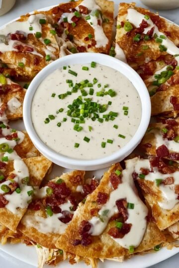Crispy Chicken Bacon Ranch Quesadillas slices arranged around a bowl of creamy ranch dipping sauce, topped with crumbled bacon and fresh green onions. A delicious cheesy appetizer and easy party snack idea perfect for quick meals or game day food.