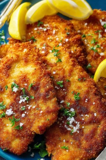 Crispy golden-brown breaded chicken schnitzel served on a blue plate, garnished with chopped parsley, coarse salt, and lemon wedges, with a fork on the side.