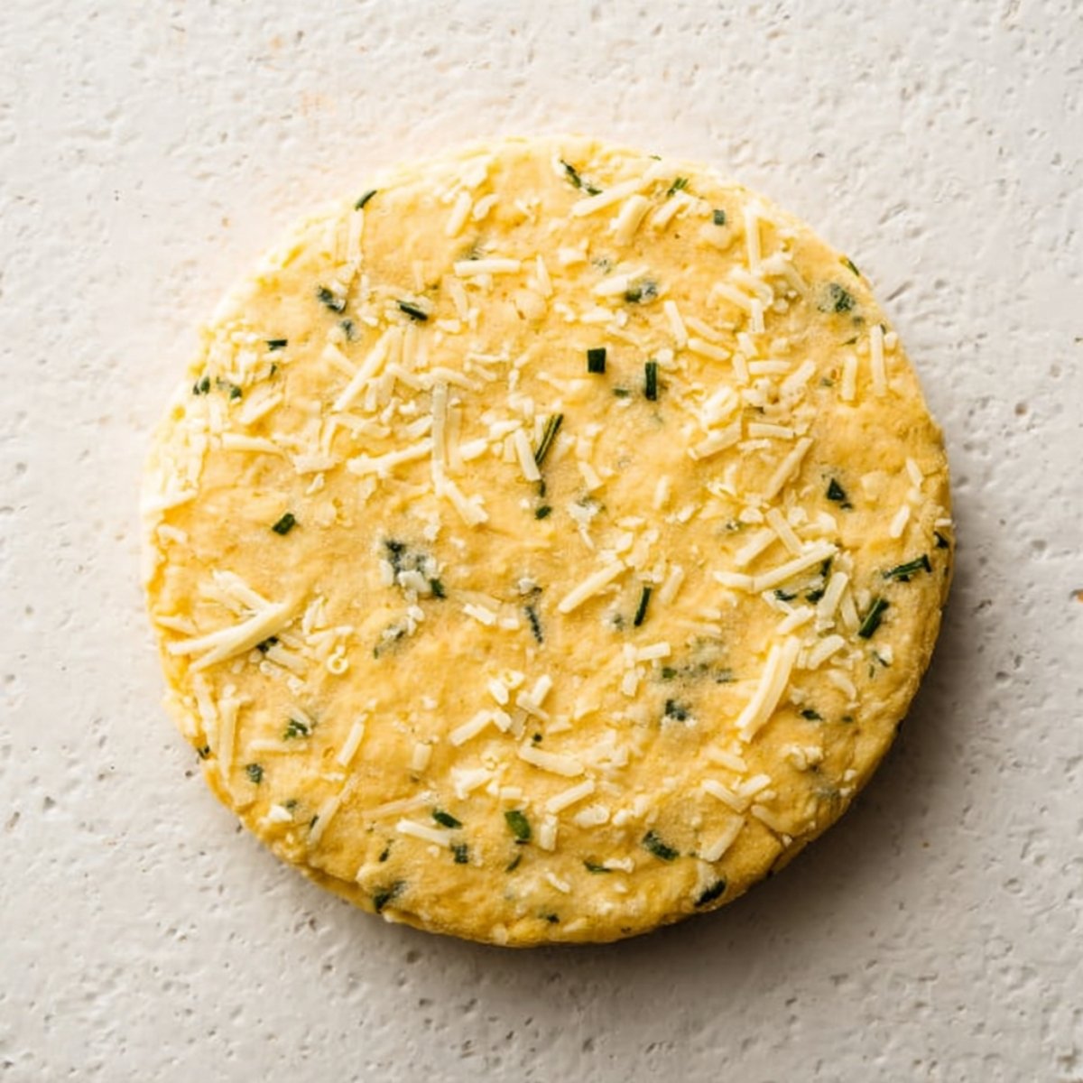 Close-up of a round disk of dough mixed with herbs and grated cheese, ready to be baked.