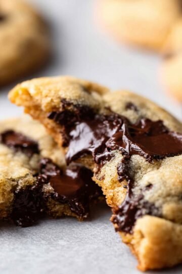 Macro shot of a Chocolate Chip Cookies broken open on a parchment-lined surface, revealing melted chocolate oozing from the center, highlighting a soft, chewy texture.