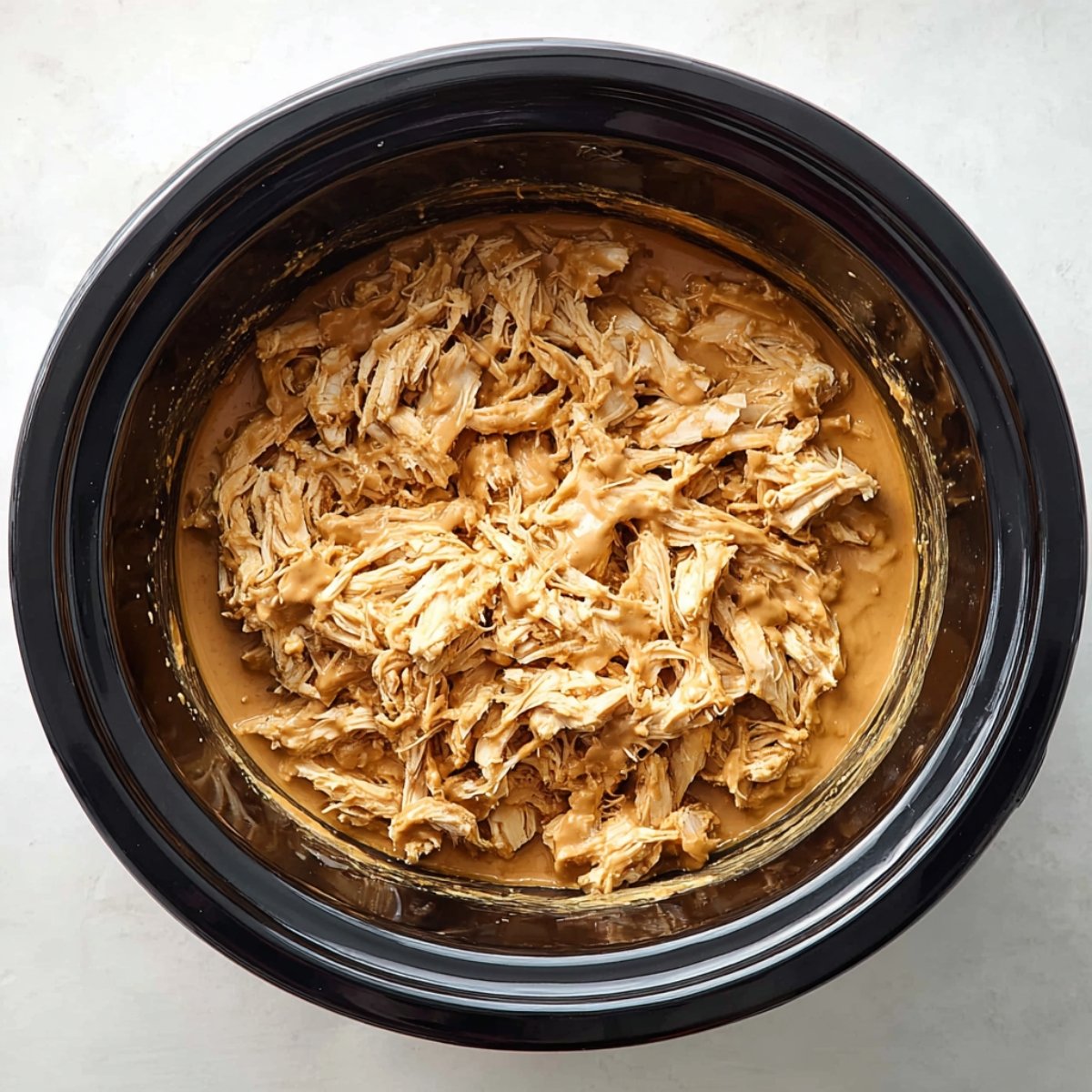 Top-down view of a black slow cooker filled with shredded chicken coated in creamy Thai peanut sauce. The chicken is fully cooked and evenly mixed with the sauce, ready to be served. Bright natural lighting, clean white countertop background, high-resolution food photography, warm and appetizing presentation.