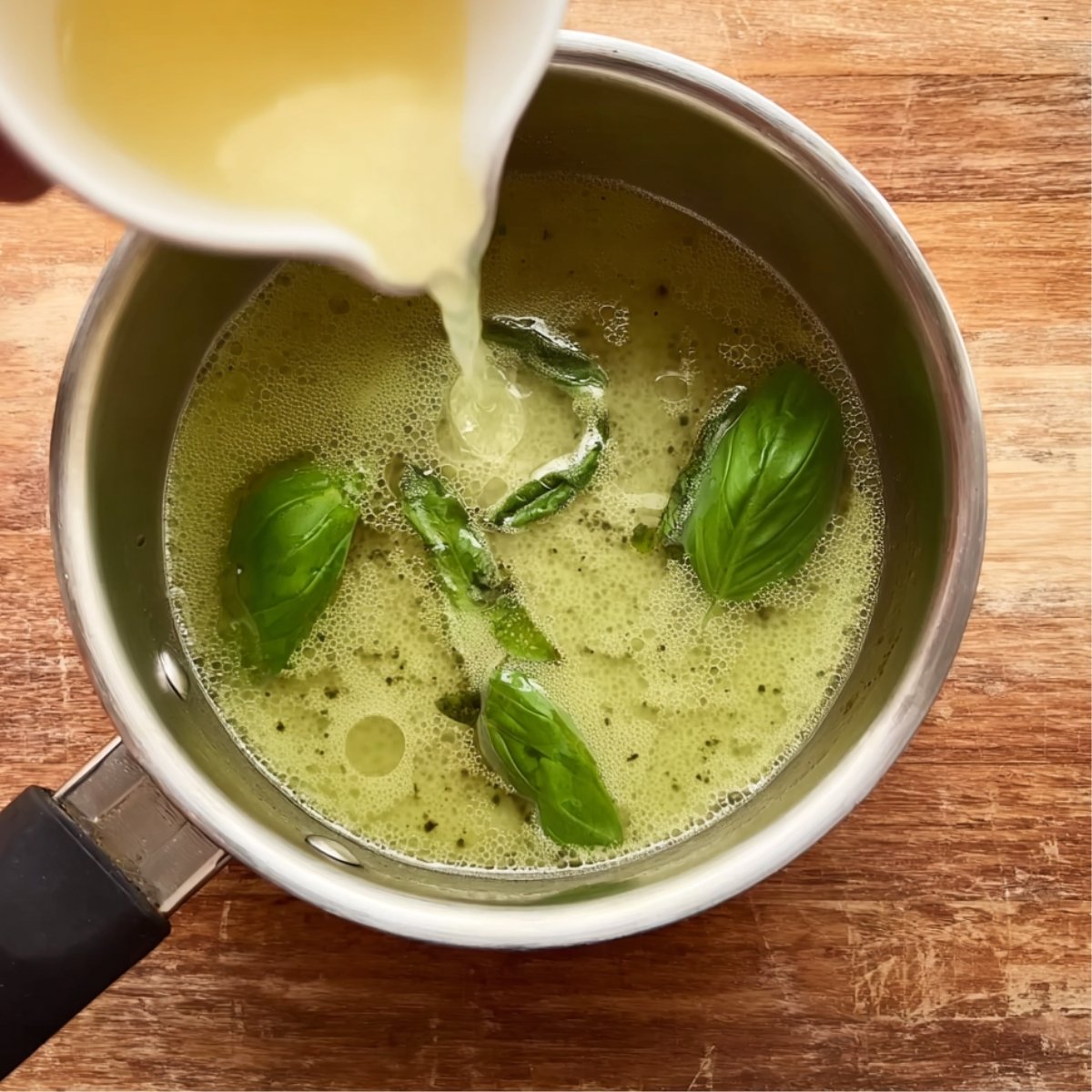 A small saucepan containing a green basil mixture as a light-colored liquid is being poured in, sitting on a wooden cutting board.