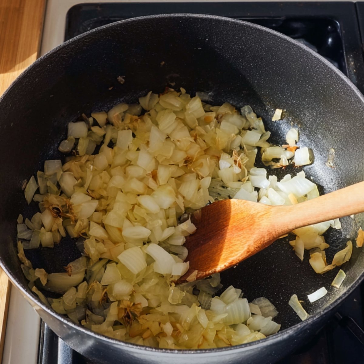 A close-up of a black pot on a stove with diced onions and garlic being sautéed with a wooden spatula, preparing the base for a curry, with a bowl of colorful spices in the background.