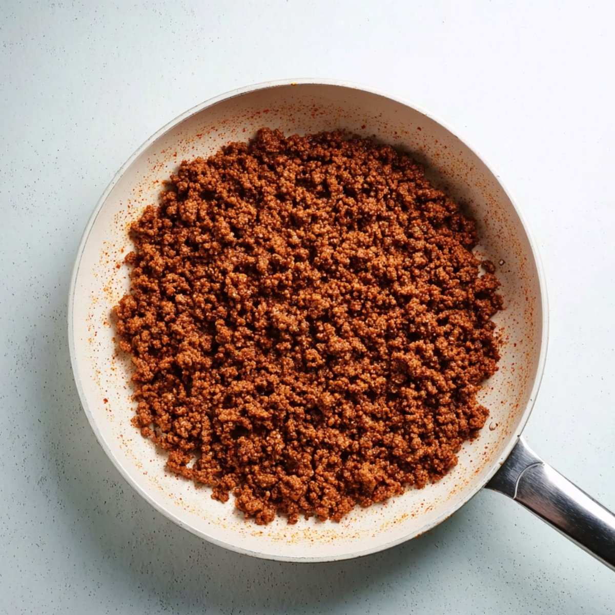 Seasoned ground beef cooking in a white skillet, browned and crumbled evenly across the pan with visible spices.