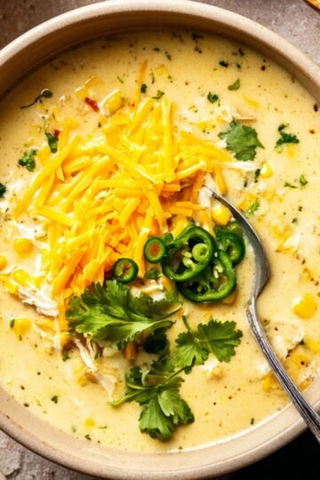 Bowl of creamy chicken poblano soup topped with shredded cheddar cheese, sliced jalapeños, and fresh cilantro leaves, with a spoon in the soup, styled for a cozy homemade meal.
