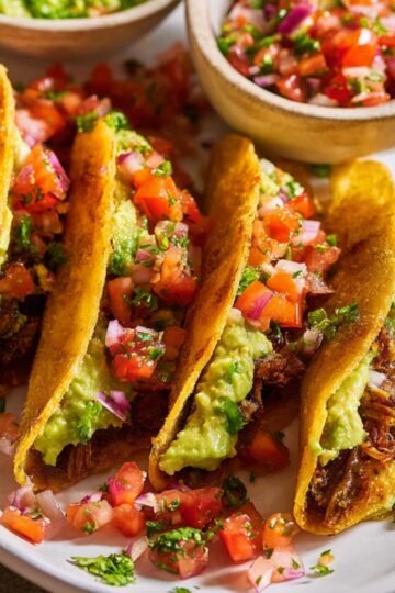 Three Crispy Ground Beef Tacos filled with shredded meat, topped with creamy guacamole and fresh pico de gallo made of diced tomatoes, onions, and herbs, served on a plate with small bowls of salsa in the background.
