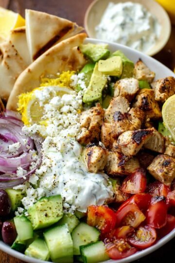 A vibrant Greek Chicken Bowls with grilled chicken chunks, diced cucumbers, cherry tomatoes, red onion slices, Kalamata olives, crumbled feta, yellow rice, and pita wedges, drizzled with creamy tzatziki sauce and garnished with lemon slices.
