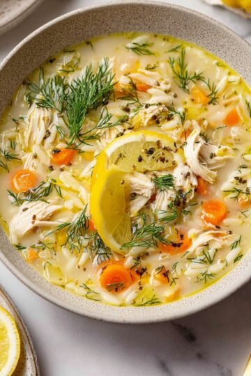 A bowl of Greek lemon chicken soup garnished with fresh dill and a slice of lemon, with a spoon resting inside and slices of bread on the side.