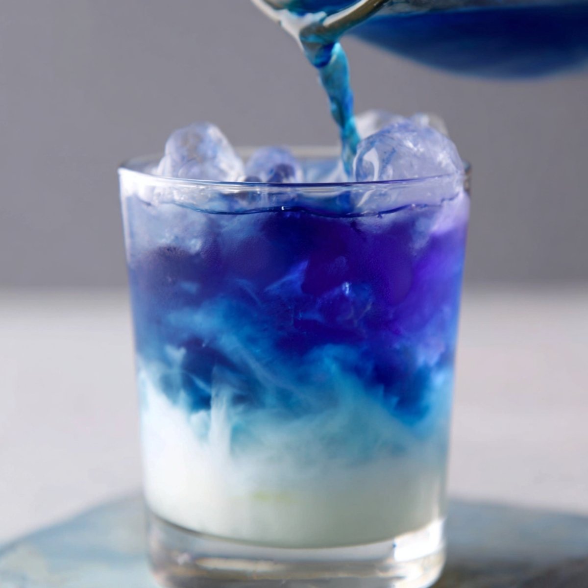 Close-up of a glass filled with ice, with vibrant blue tea being poured on top, creating a layered ombre effect over a white base.