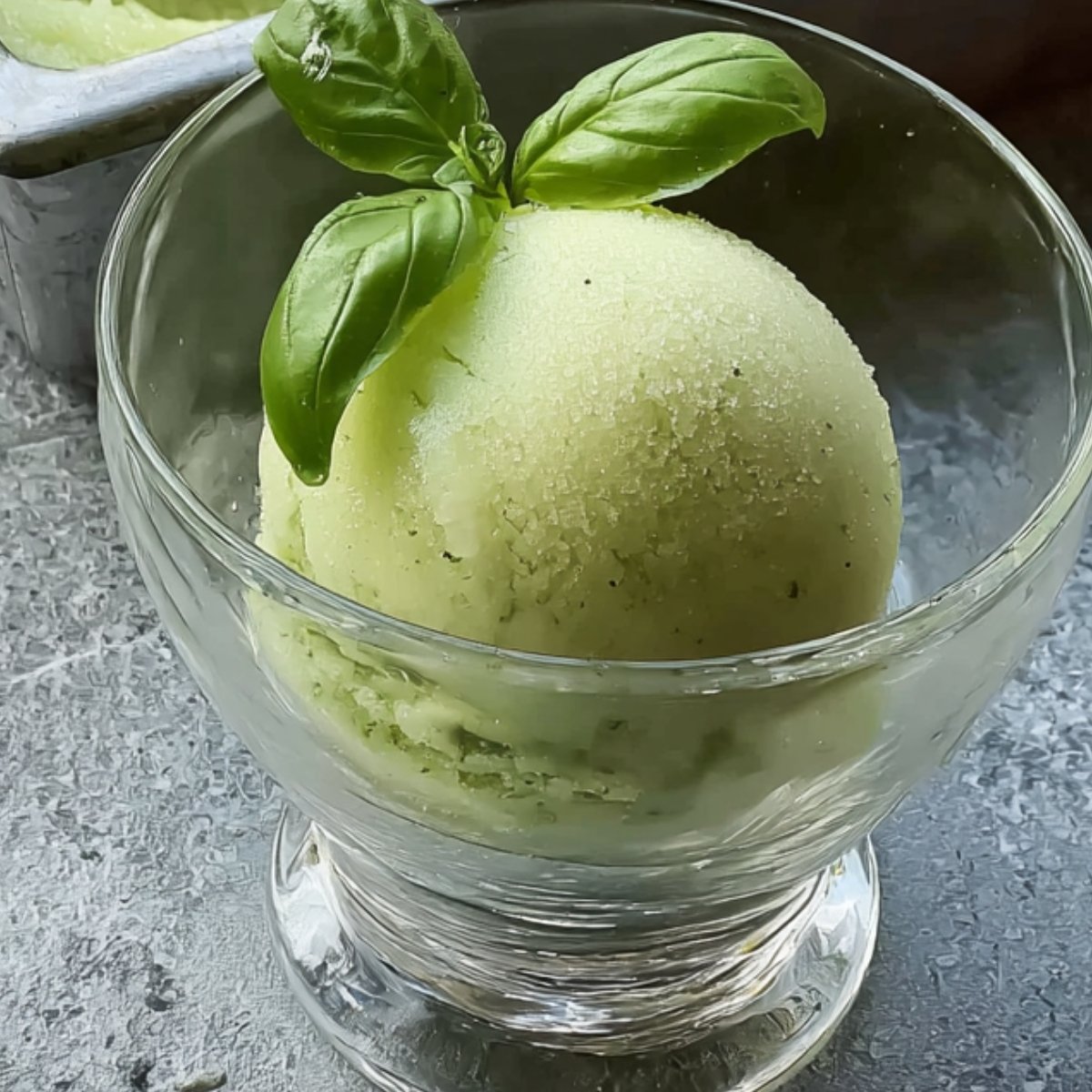 A glass dessert bowl holding a smooth scoop of light green Lemon Basil Sorbet, garnished with fresh basil leaves, placed on a chilled surface.
