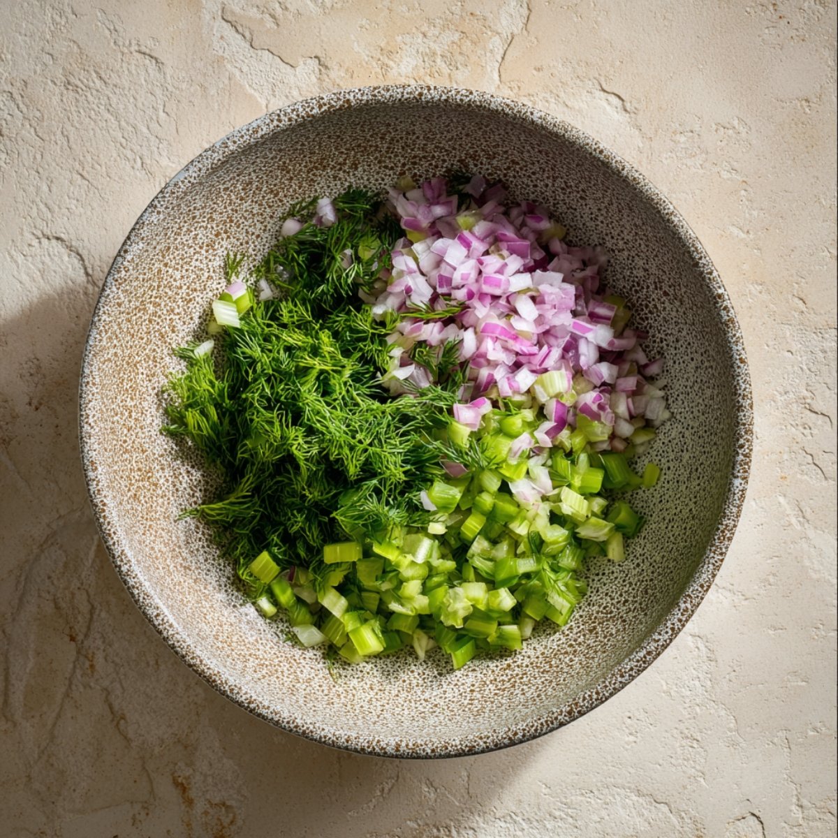 Bowl with chopped red onion, diced celery, and fresh dill arranged together on a light textured surface.