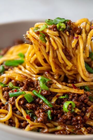 Close-up of a bowl of Mongolian Ground Beef Noodles, with tender noodles coated in a rich dark sauce, mixed with ground beef, garnished with chopped green onions and chili flakes. A fork lifts a portion of noodles, showing the glossy, saucy texture, on a wooden surface background.