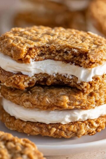 Stack of oatmeal creme pies on a white plate, one showing a bite taken out to reveal the fluffy white cream filling. The golden-brown oatmeal cookies have a textured surface, with additional cookies and a glass of milk softly blurred in the background.
