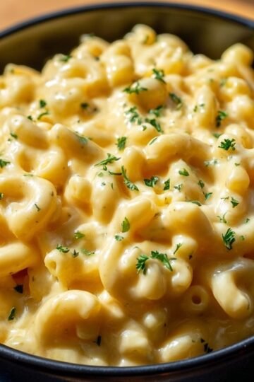 Close-up of creamy one-pot macaroni and cheese in a black bowl, elbow pasta coated in smooth, melted cheddar cheese sauce, garnished with small parsley flakes, bright natural lighting, realistic food photography, warm and comforting presentation.