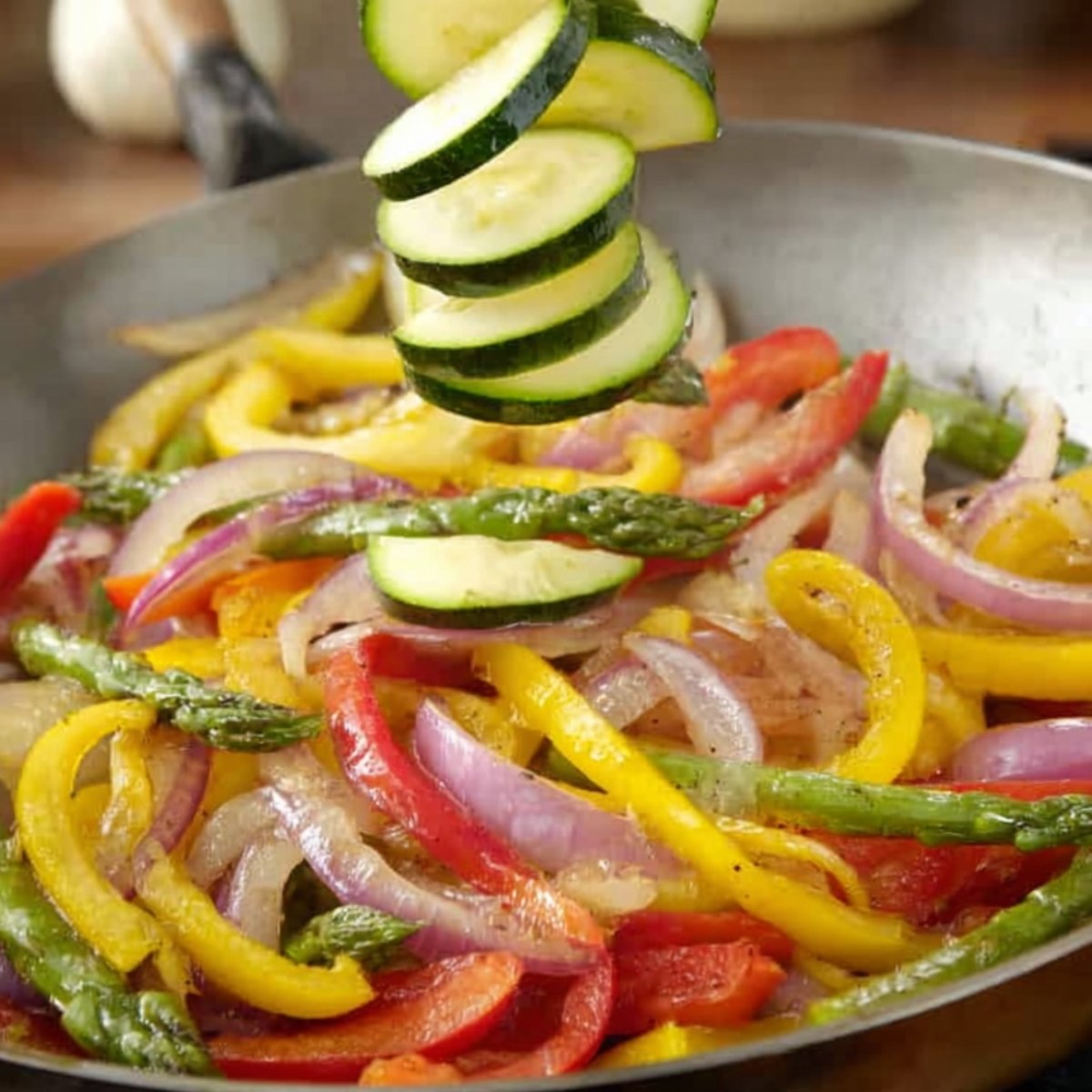 Sliced zucchini being added to a pan of sautéed vegetables including bell peppers, red onions, and asparagus.