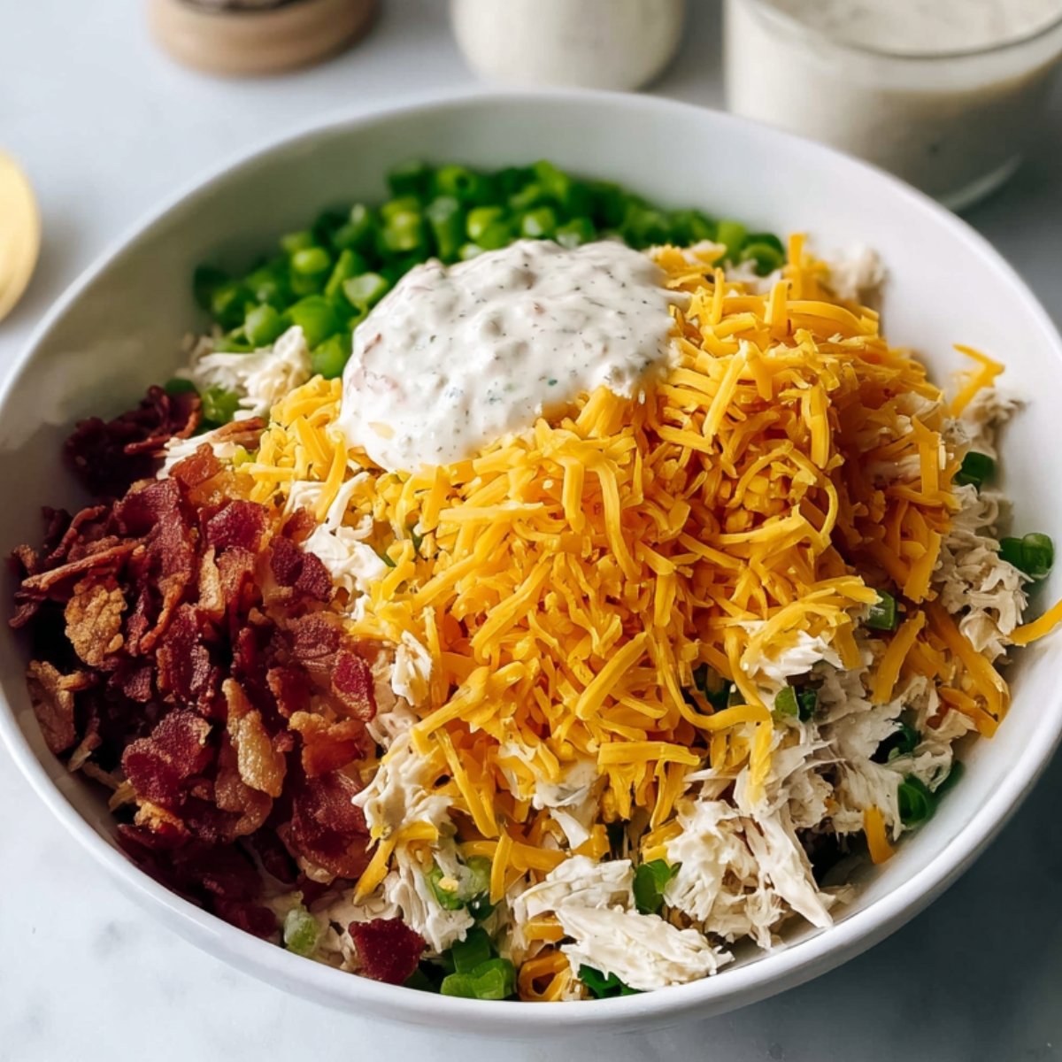 A white bowl filled with prepared ingredients for a chicken salad or casserole, including shredded cooked chicken, chopped green onions, crispy bacon bits, shredded cheddar cheese, and a dollop of creamy dressing on top. The ingredients are arranged in sections, ready to be mixed.