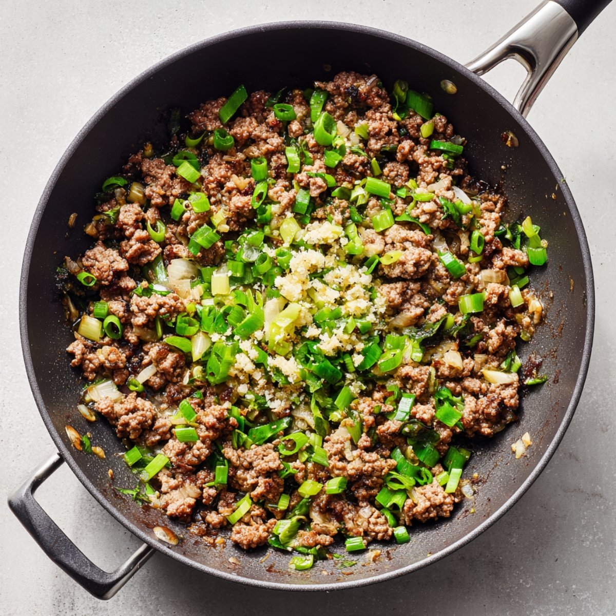 Top-down view of a skillet with cooked ground beef, chopped scallions, and minced garlic, showing a sizzling, savory cooking-in-progress scene with vibrant green vegetables contrasting the browned beef.