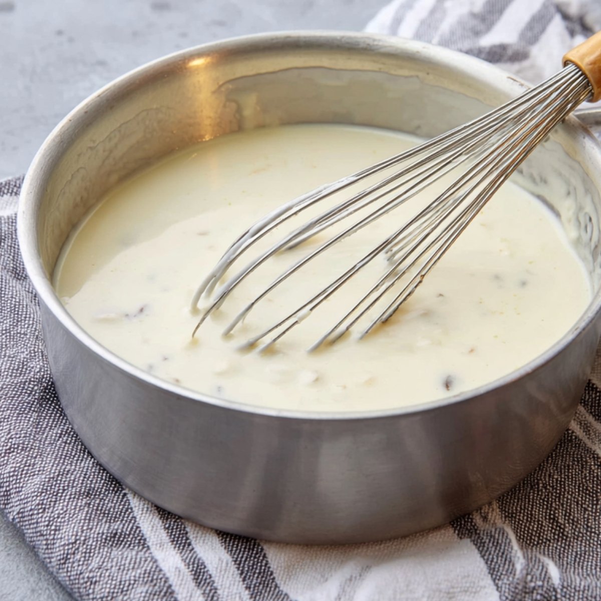 Close-up of a saucepan with creamy white sauce being whisked, smooth texture with small bits, set on a striped cloth, natural lighting, high-resolution food preparation.