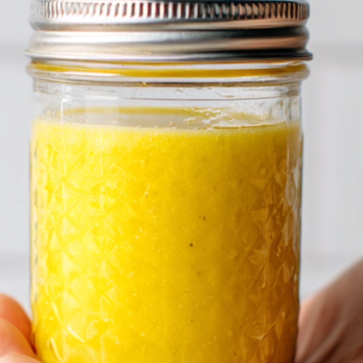 A close-up of a mason jar filled with bright yellow salad dressing, held in hand. The dressing appears smooth and creamy, ready to be poured over a salad.