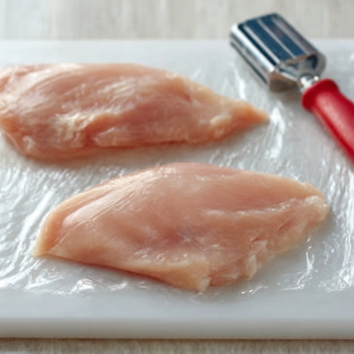Two raw chicken breasts placed on a sheet of plastic wrap on a cutting board, next to a metal meat tenderizer with a red handle, ready to be pounded for even thickness.