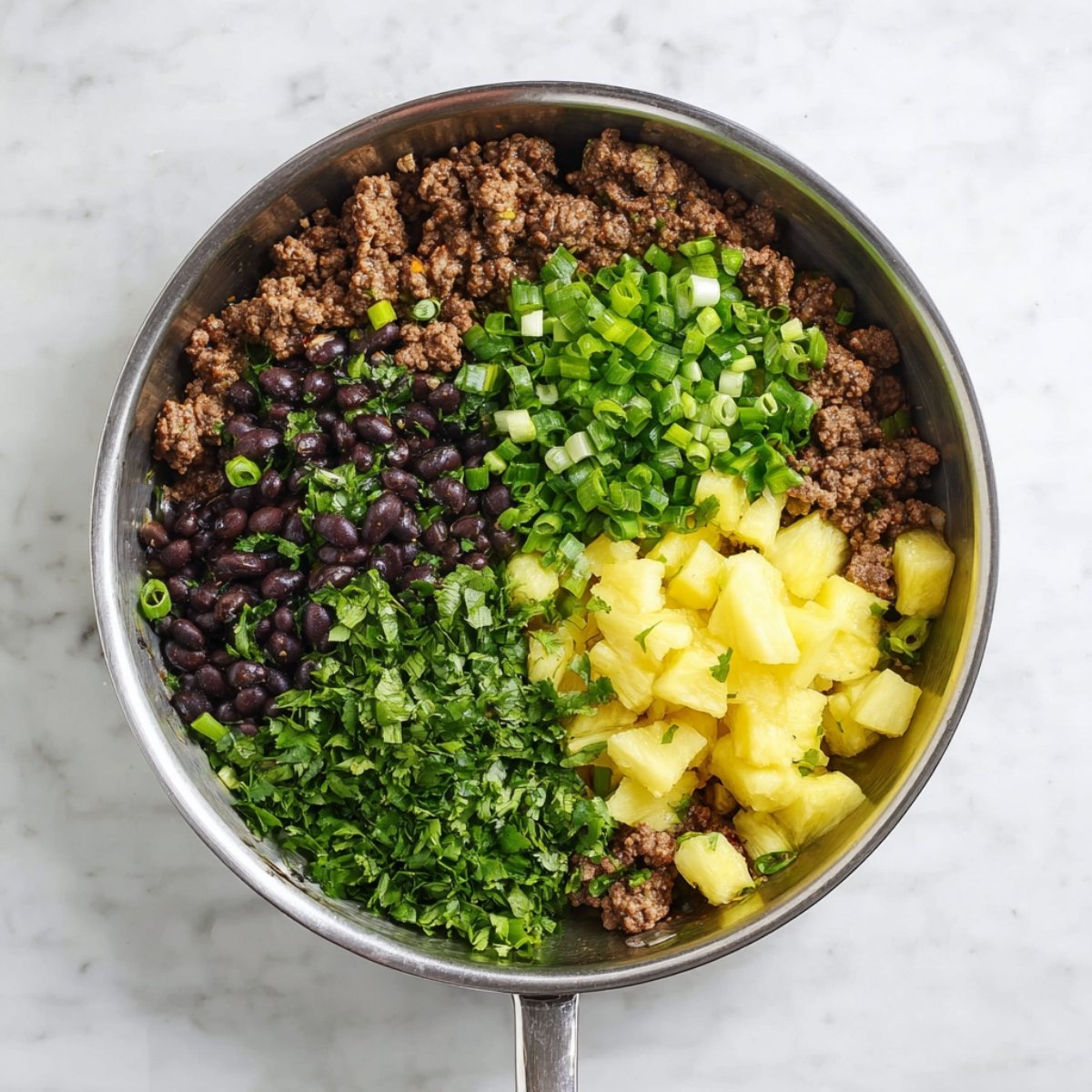 Top-down view of a stainless steel skillet with cooked ground beef, black beans, diced pineapple, chopped green onions, and fresh cilantro, ingredients not yet mixed, on a white countertop, bright natural lighting, fresh and vibrant presentation.
