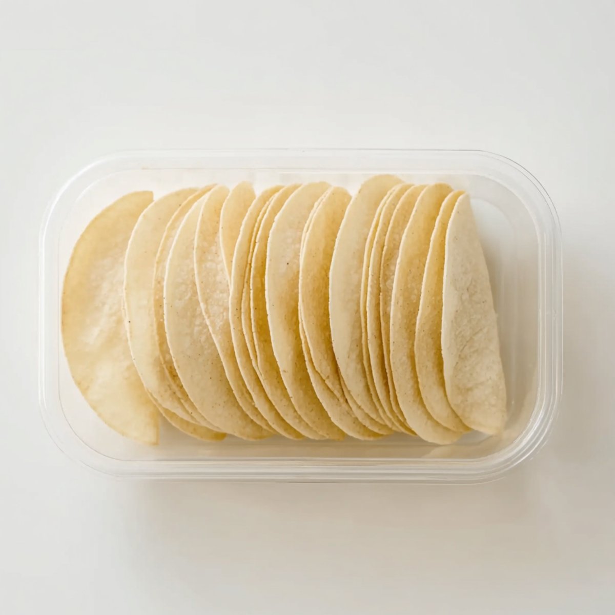 A clear plastic container holding a stack of small, soft corn tortillas neatly arranged side by side.