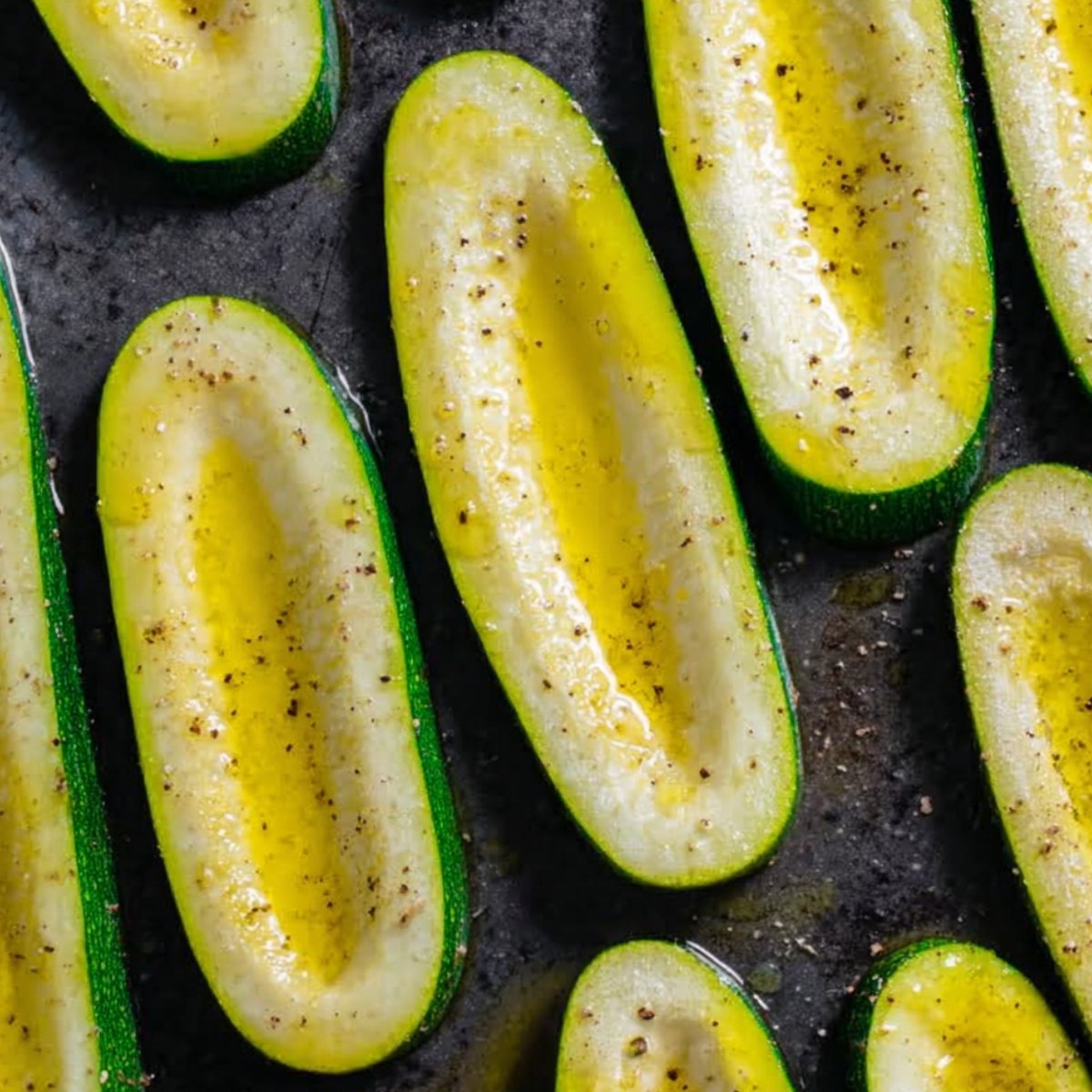 Halved green zucchini arranged on a baking tray, drizzled with olive oil and sprinkled with black pepper, ready for roasting.