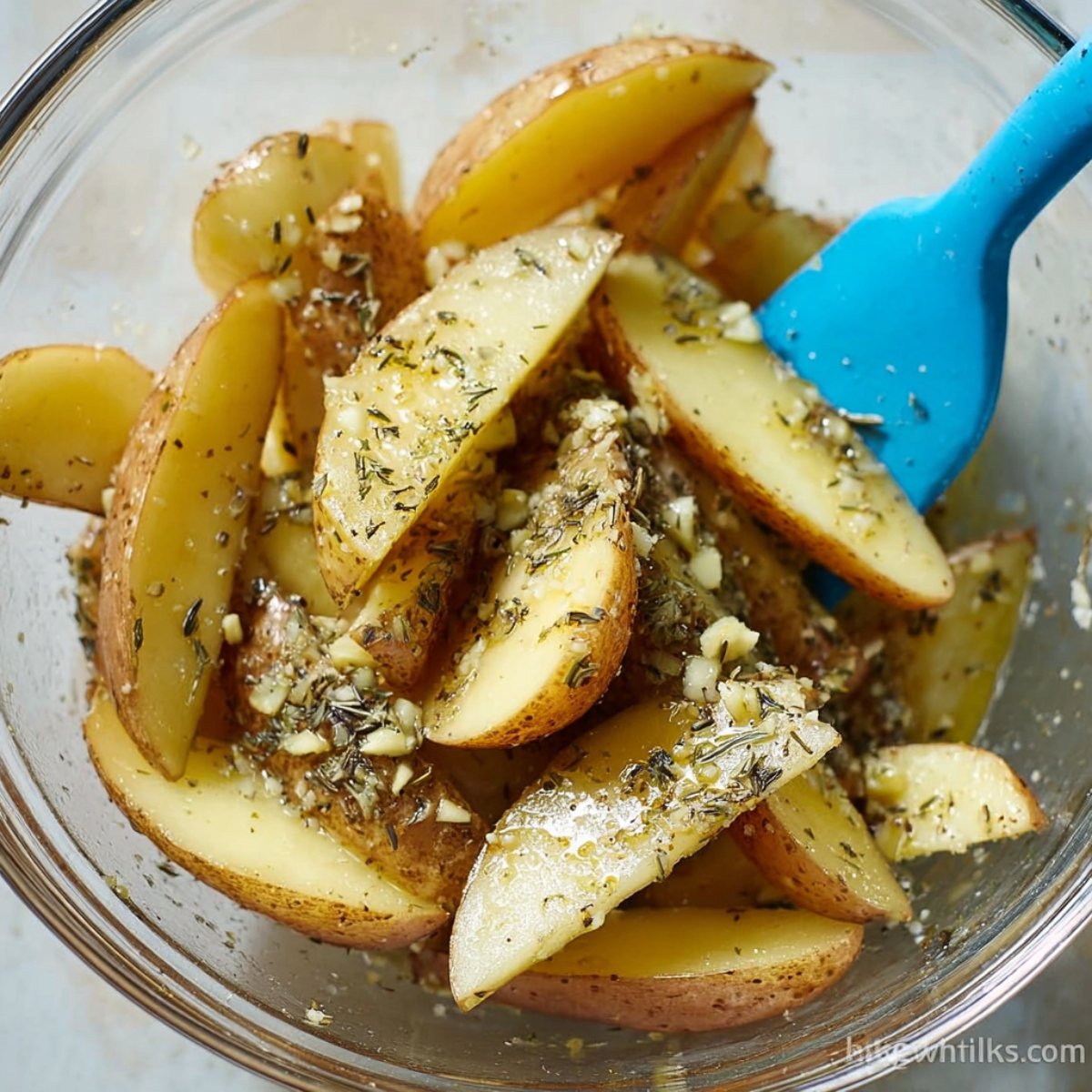 Close-up of potato wedges in a glass mixing bowl, coated with minced garlic, olive oil, and dried herbs, with a blue silicone spatula for tossing, ready for baking.