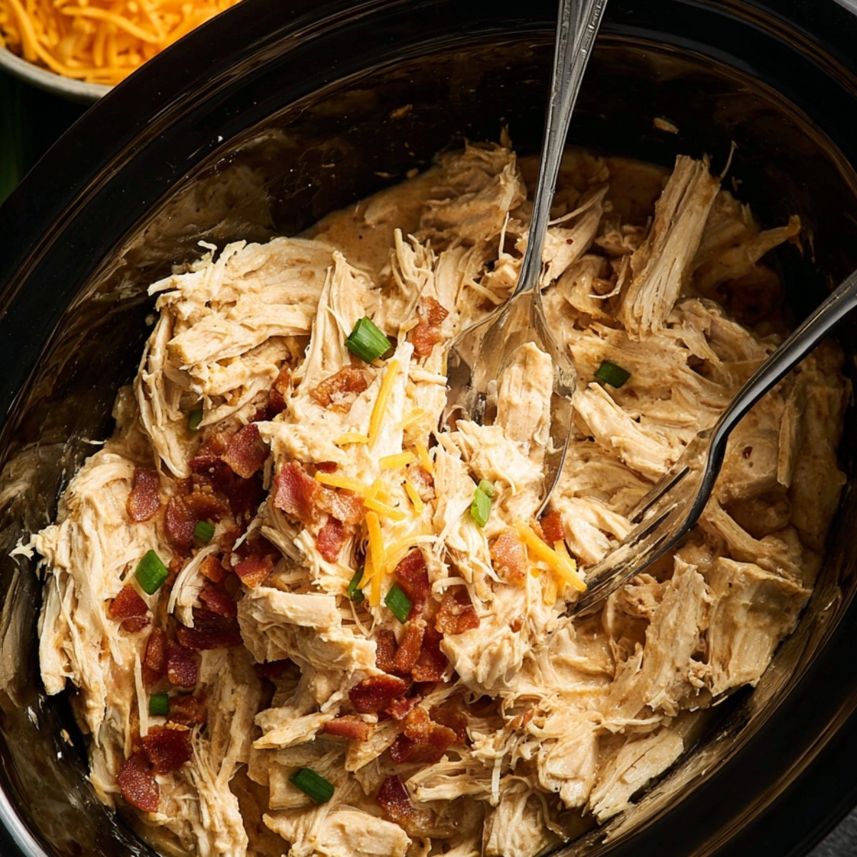 Top-down view of shredded slow cooker chicken being mixed with cream cheese in a black slow cooker, showing creamy sauce beginning to coat the chicken. A bowl of shredded cheddar is visible in the background along with green onions.