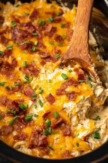 Close-up of shredded Slow Cooker Crack Chicken mixed with creamy sauce, topped with melted cheddar cheese, crispy bacon bits, and chopped green onions in a black slow cooker. A wooden spoon scoops a portion of the cheesy mixture.