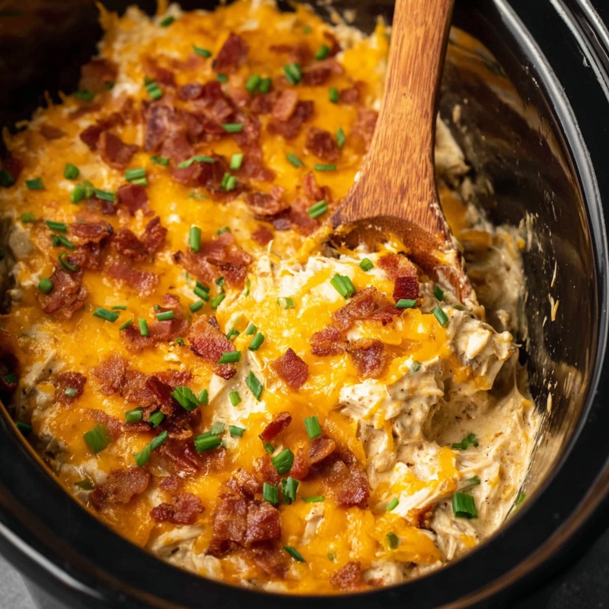 Close-up of shredded Slow Cooker Crack Chicken mixed with creamy sauce, topped with melted cheddar cheese, crispy bacon bits, and chopped green onions in a black slow cooker. A wooden spoon scoops a portion of the cheesy mixture.