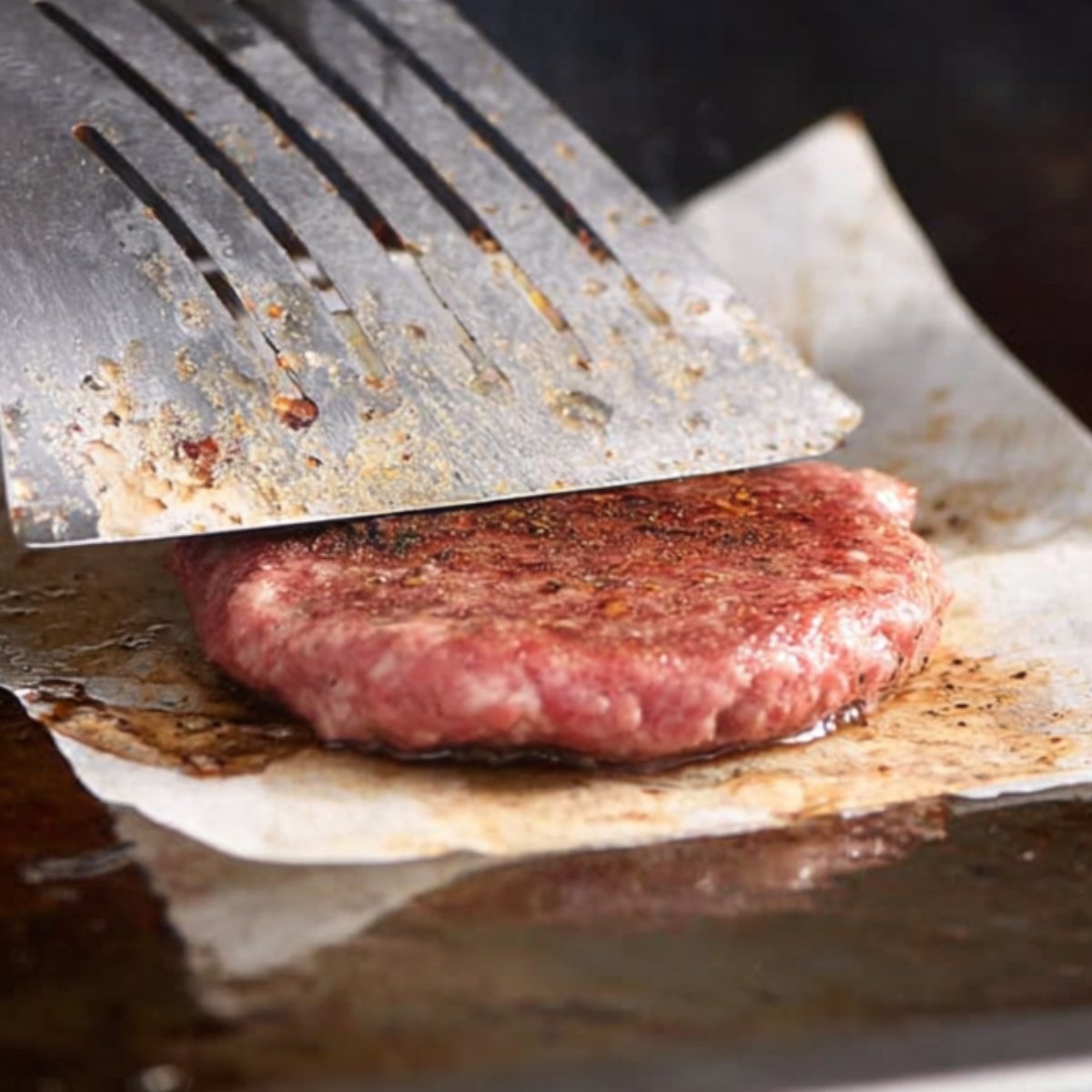Raw ground beef patty being pressed on a hot griddle with a metal spatula, using parchment paper, showing the action of smashing the burger for a crispy crust.