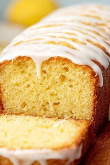 Close-up of a sliced Starbucks Copycat Lemon Loaf on a wooden board, topped with white glaze drizzled over the golden cake. The cake has a moist, soft yellow crumb, with fresh lemon slices placed nearby.