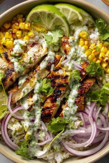 Top-down view of a completed street corn chicken bowl with grilled chicken slices, charred corn, white rice, sliced red onions, fresh cilantro, lime wedges, and creamy herb sauce drizzled on top, presented in a beige bowl on a gray surface.