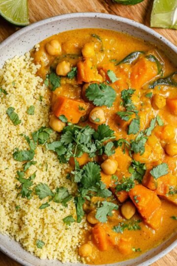 A top-down view of a bowl filled with Sweet Potato and Chickpea Curry curry served alongside fluffy couscous, garnished with fresh cilantro and lime wedges, placed on a wooden board with a cloth napkin.