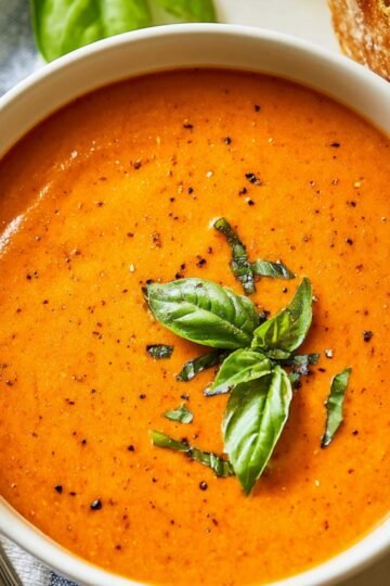Creamy tomato bisque soup in a white bowl, garnished with fresh basil leaves, placed on a striped cloth with a piece of crusty bread partially visible beside it.