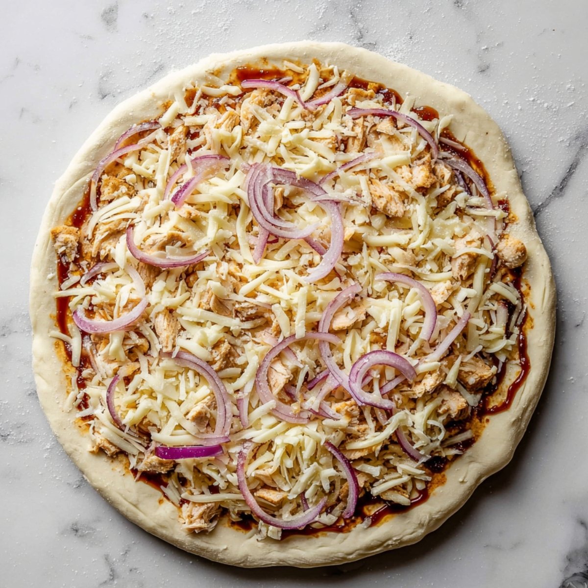Unbaked BBQ chicken pizza on a marble countertop, topped with shredded mozzarella and cheddar cheese, shredded cooked chicken, and thinly sliced red onions. The pizza has a hand-stretched crust with a slightly raised edge, and the toppings are evenly distributed across the surface, ready to be baked.
