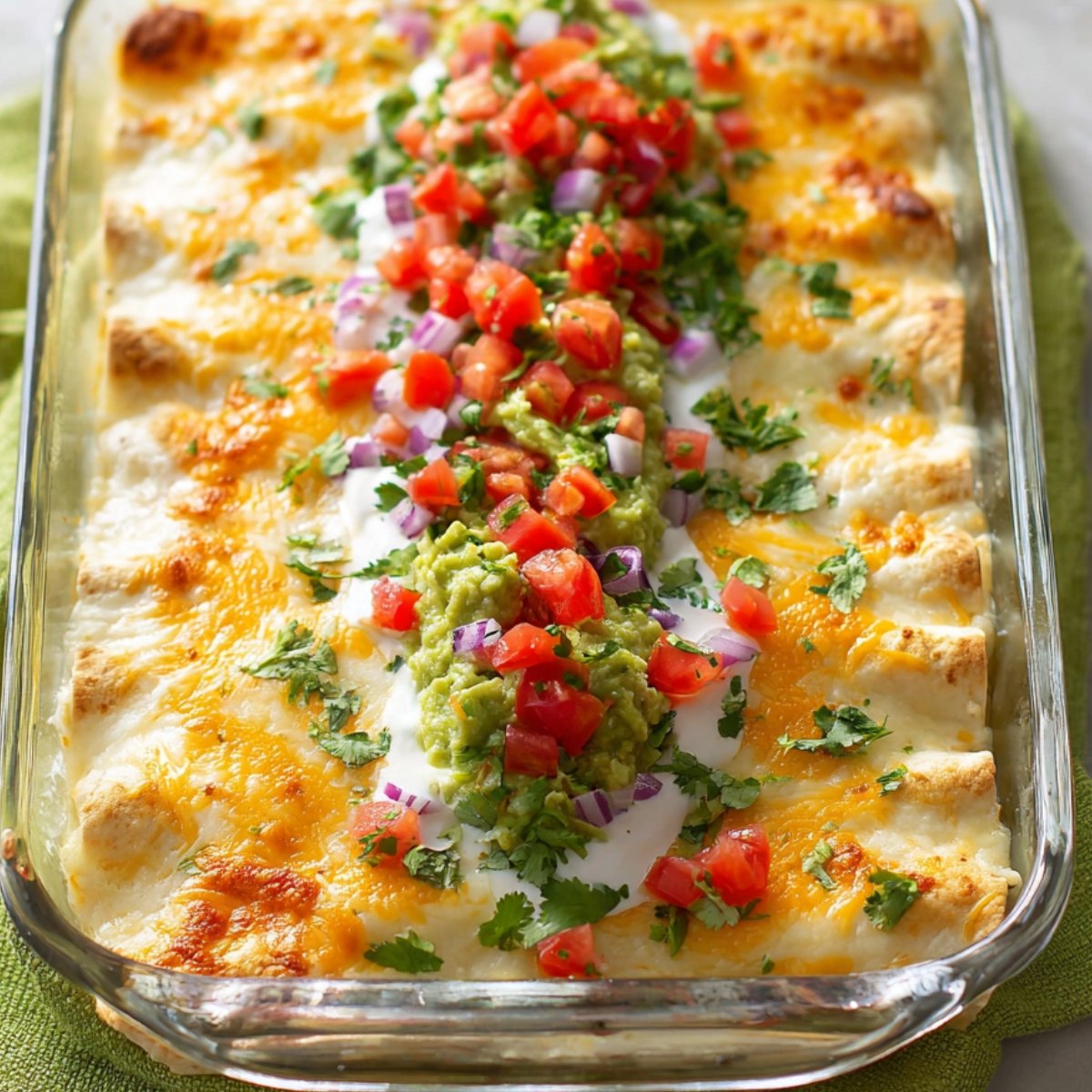 Glass baking dish with baked white chicken enchiladas, topped with melted cheese, garnished with diced tomatoes, red onions, guacamole, sour cream, and fresh cilantro, set on a green cloth, bright natural light.