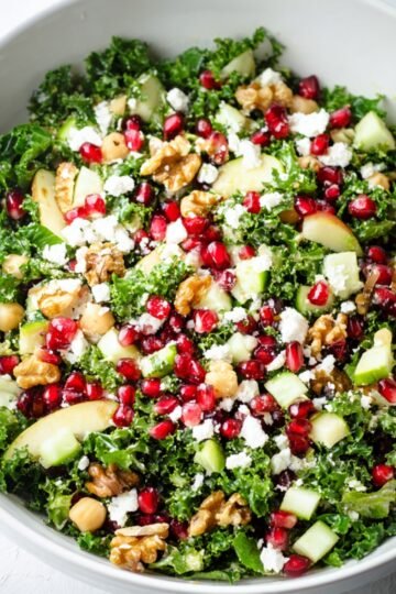 A large white bowl filled with a vibrant Winter Kale Salad. The salad contains chopped kale, pomegranate seeds, chickpeas, sliced apples, crumbled feta cheese, and walnut pieces, all mixed together.
