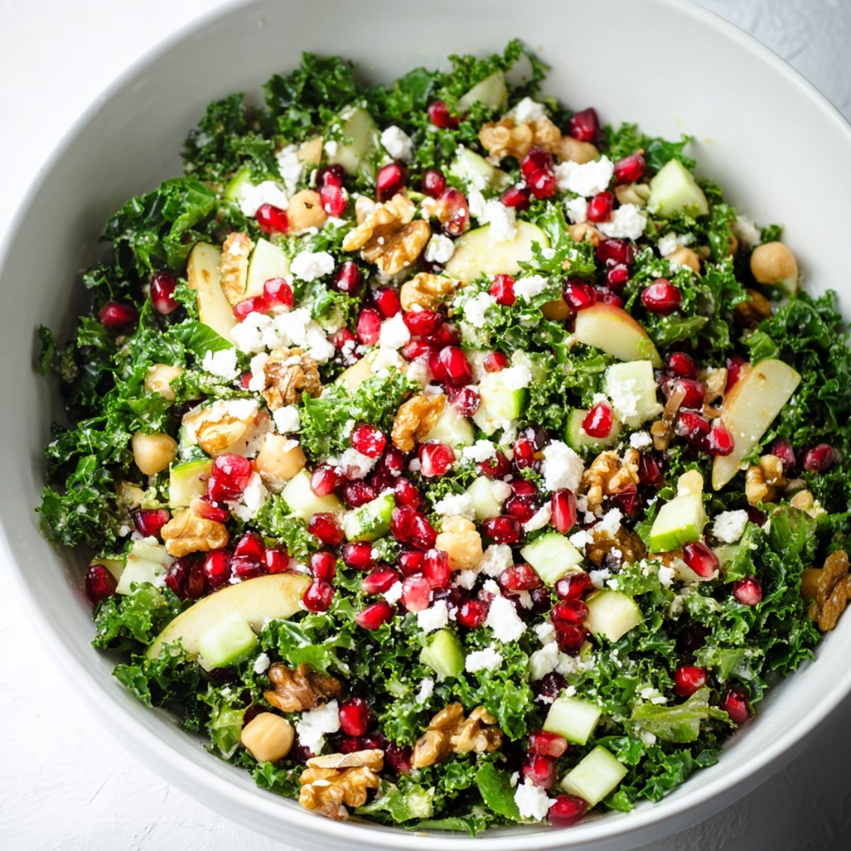 A large white bowl filled with a vibrant Winter Kale Salad. The salad contains chopped kale, pomegranate seeds, chickpeas, sliced apples, crumbled feta cheese, and walnut pieces, all mixed together.