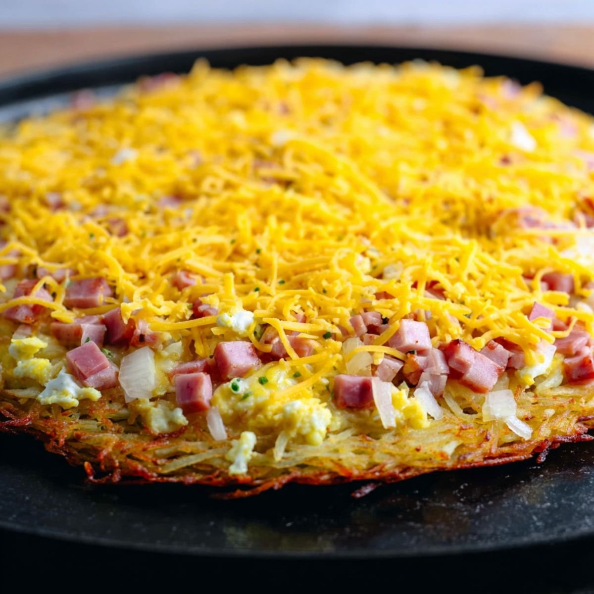 Close-up of a breakfast pizza on a black pan with a hash brown crust, topped with diced ham, scrambled eggs, and a generous layer of shredded cheddar cheese.