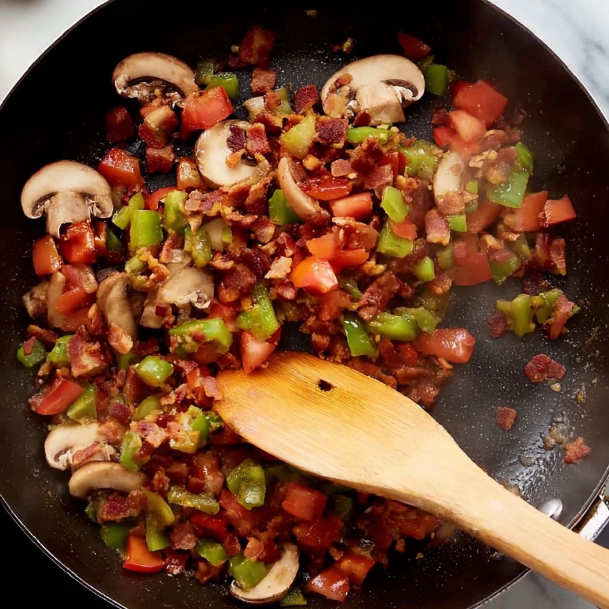 Overhead view of a sauté pan with diced tomatoes, green bell peppers, mushrooms, and crispy bacon being stirred with a wooden spatula.