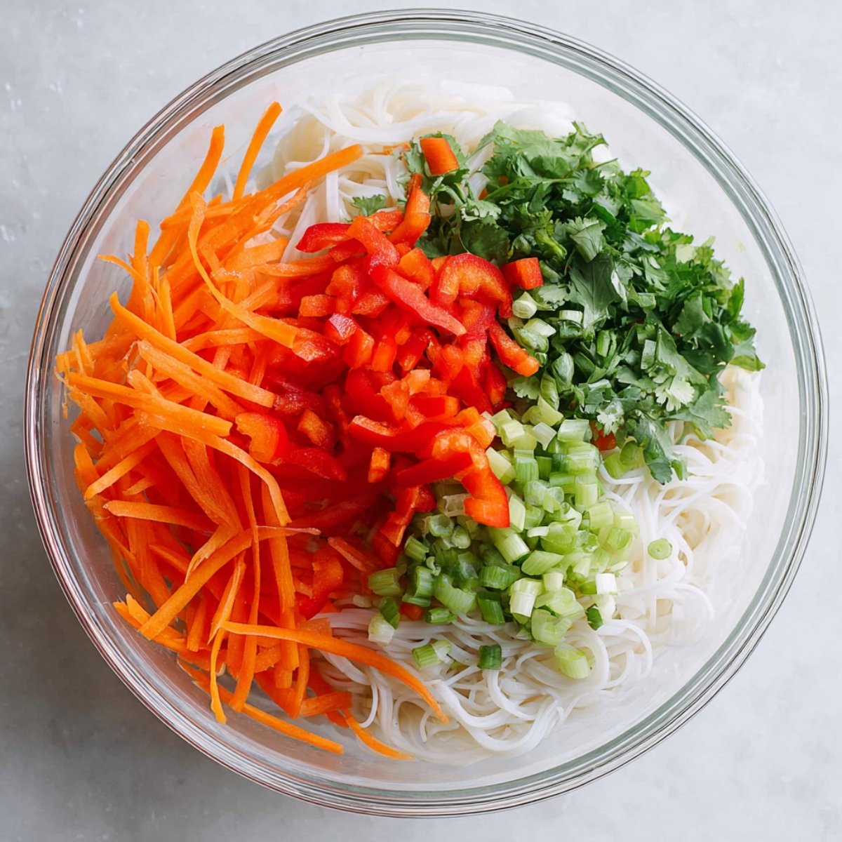 Glass bowl of rice noodles topped with shredded carrots, chopped red bell peppers, green onions, and fresh cilantro.