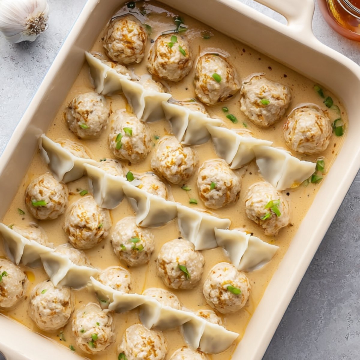 Raw meatballs and folded dumplings arranged neatly in a baking dish with a creamy sauce